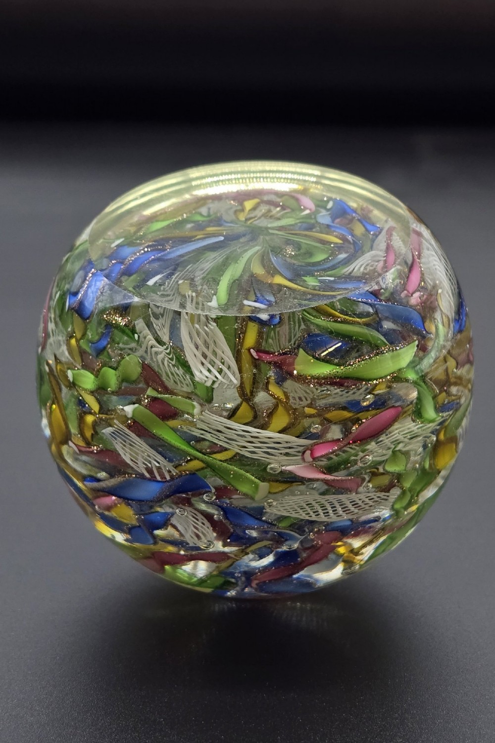 A clear spherical glass paperweight filled with an intricate mix of colourful cane fragments, white filigree ribbons, and glittering gold aventurine flecks. View V