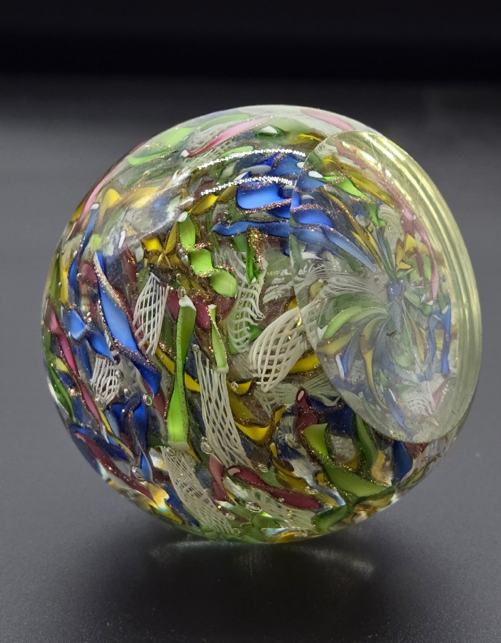 A clear spherical glass paperweight filled with an intricate mix of colourful cane fragments, white filigree ribbons, and glittering gold aventurine flecks. View VI