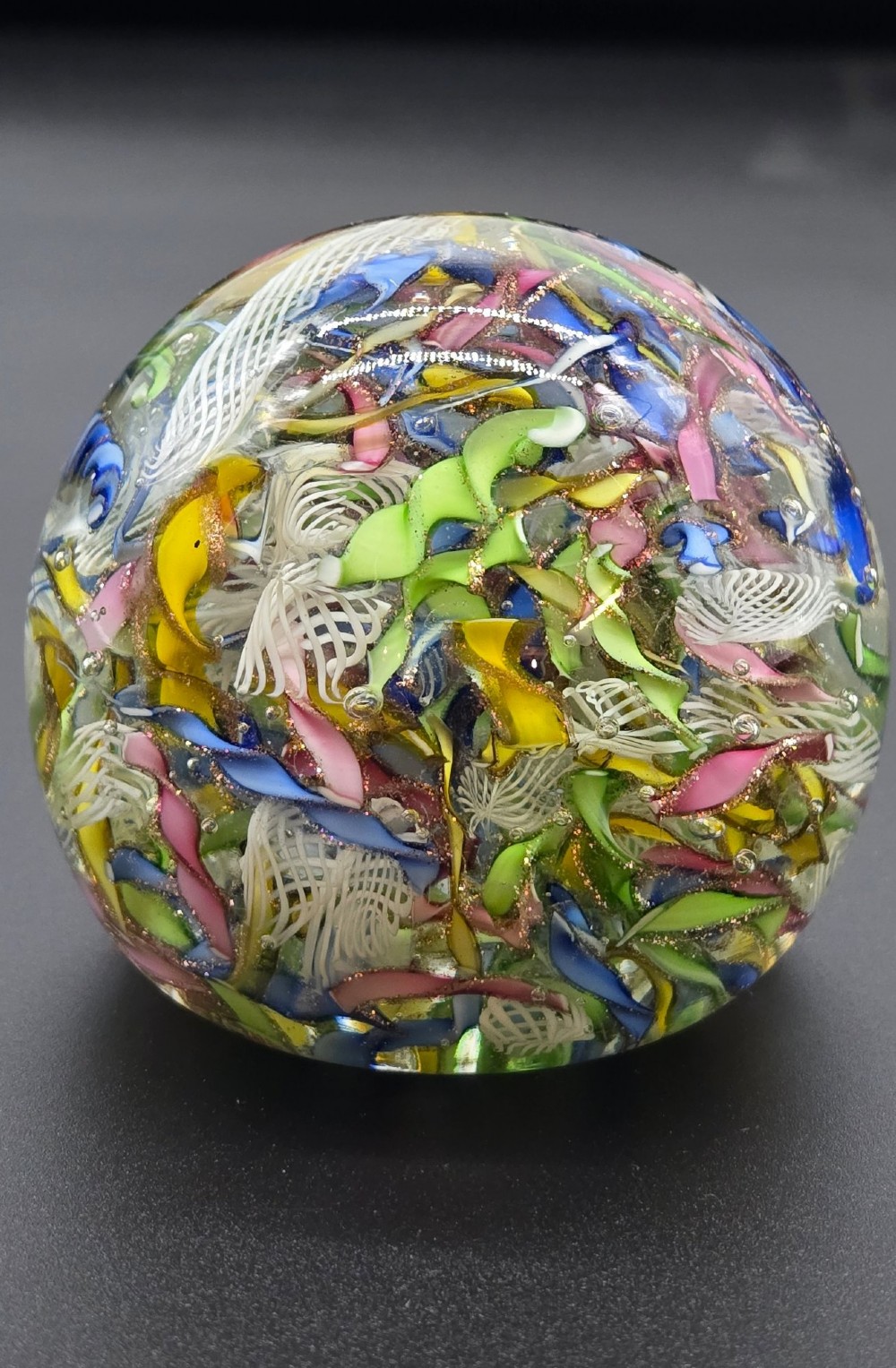 A clear spherical glass paperweight filled with an intricate mix of colourful cane fragments, white filigree ribbons, and glittering gold aventurine flecks. View VII