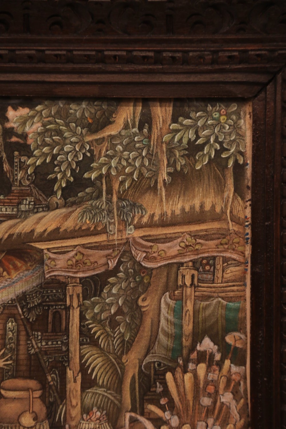 A detailed Balinese painting depicting figures dancing and playing gamelan instruments within a lush landscape, set inside a carved dark wood frame. View II