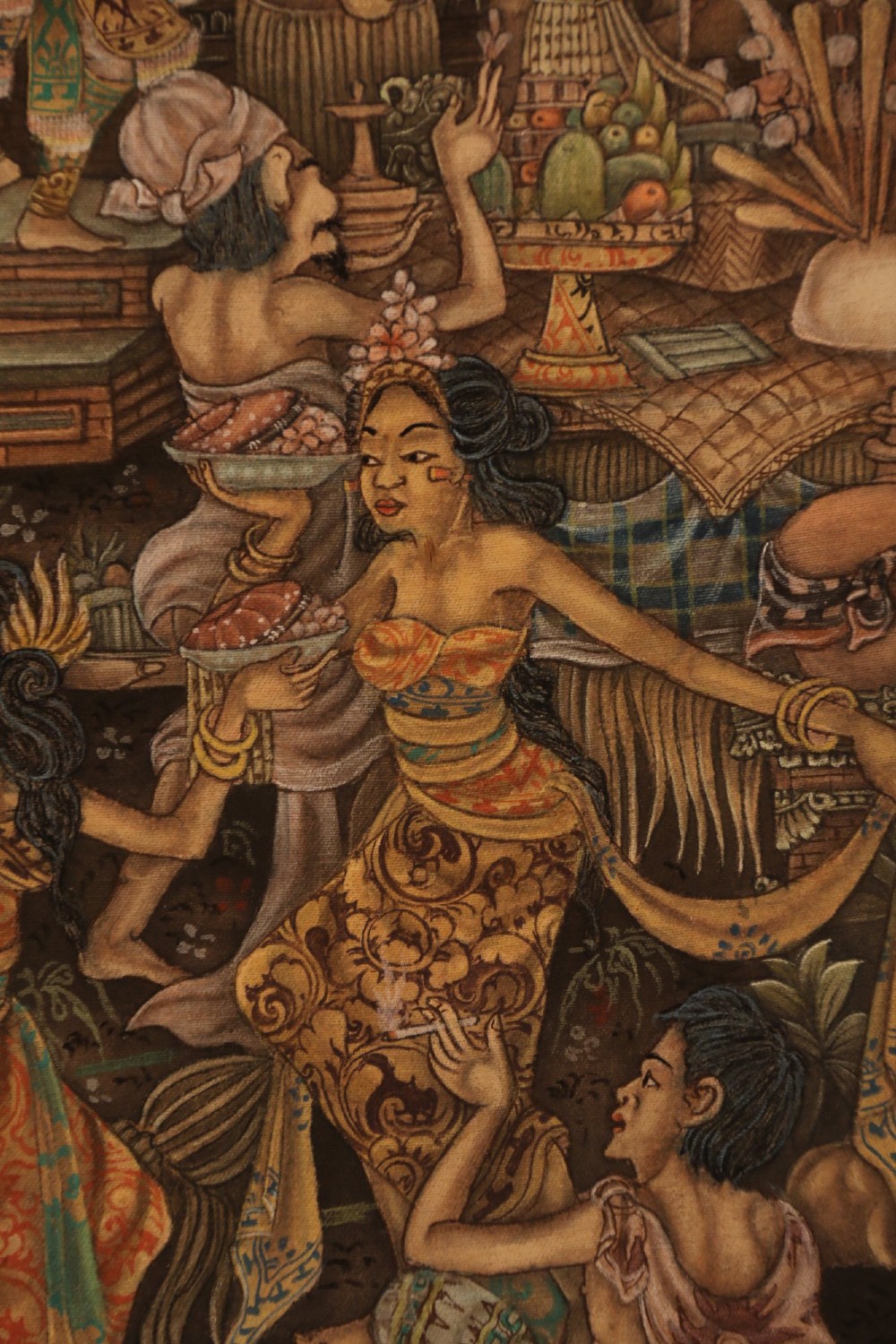 A detailed Balinese painting depicting figures dancing and playing gamelan instruments within a lush landscape, set inside a carved dark wood frame. View III