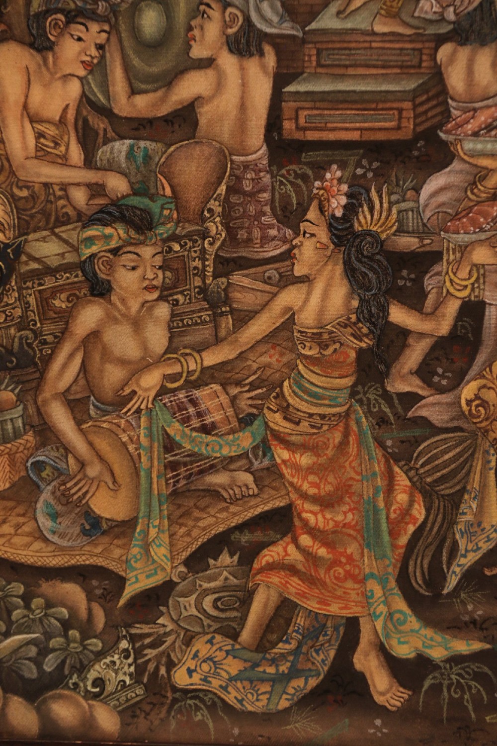 A detailed Balinese painting depicting figures dancing and playing gamelan instruments within a lush landscape, set inside a carved dark wood frame. View V
