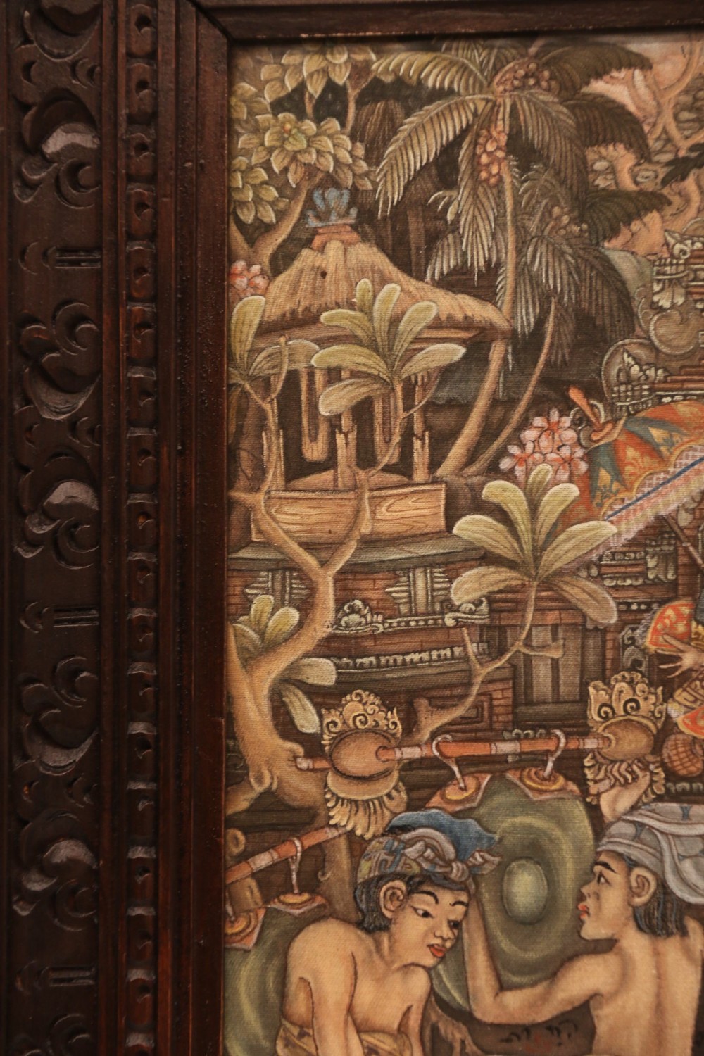 A detailed Balinese painting depicting figures dancing and playing gamelan instruments within a lush landscape, set inside a carved dark wood frame. View VIII