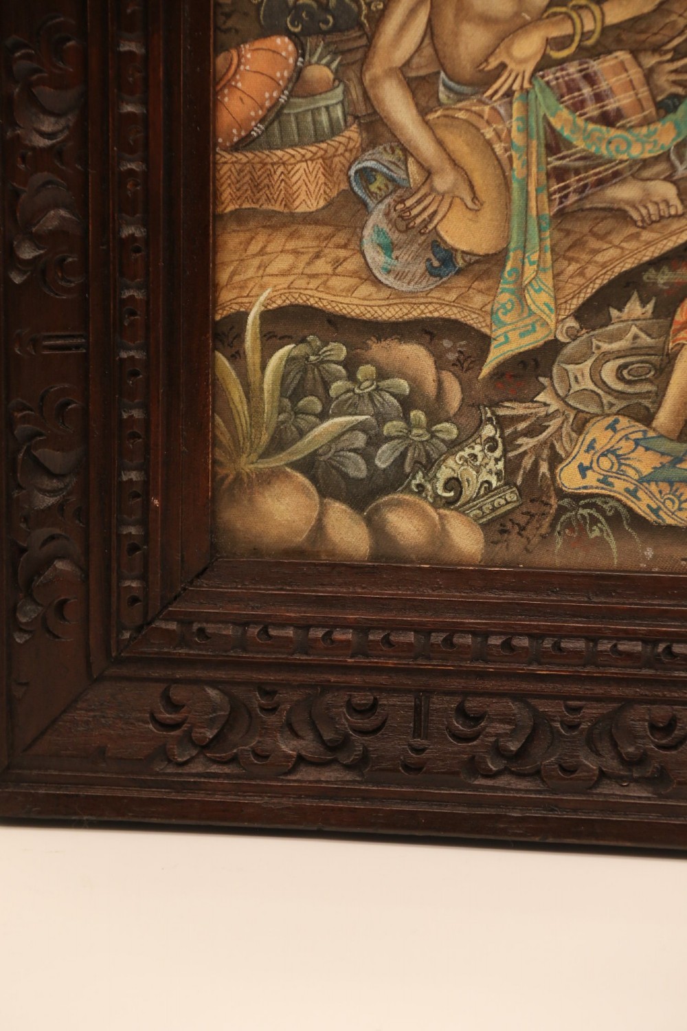 A detailed Balinese painting depicting figures dancing and playing gamelan instruments within a lush landscape, set inside a carved dark wood frame. View X