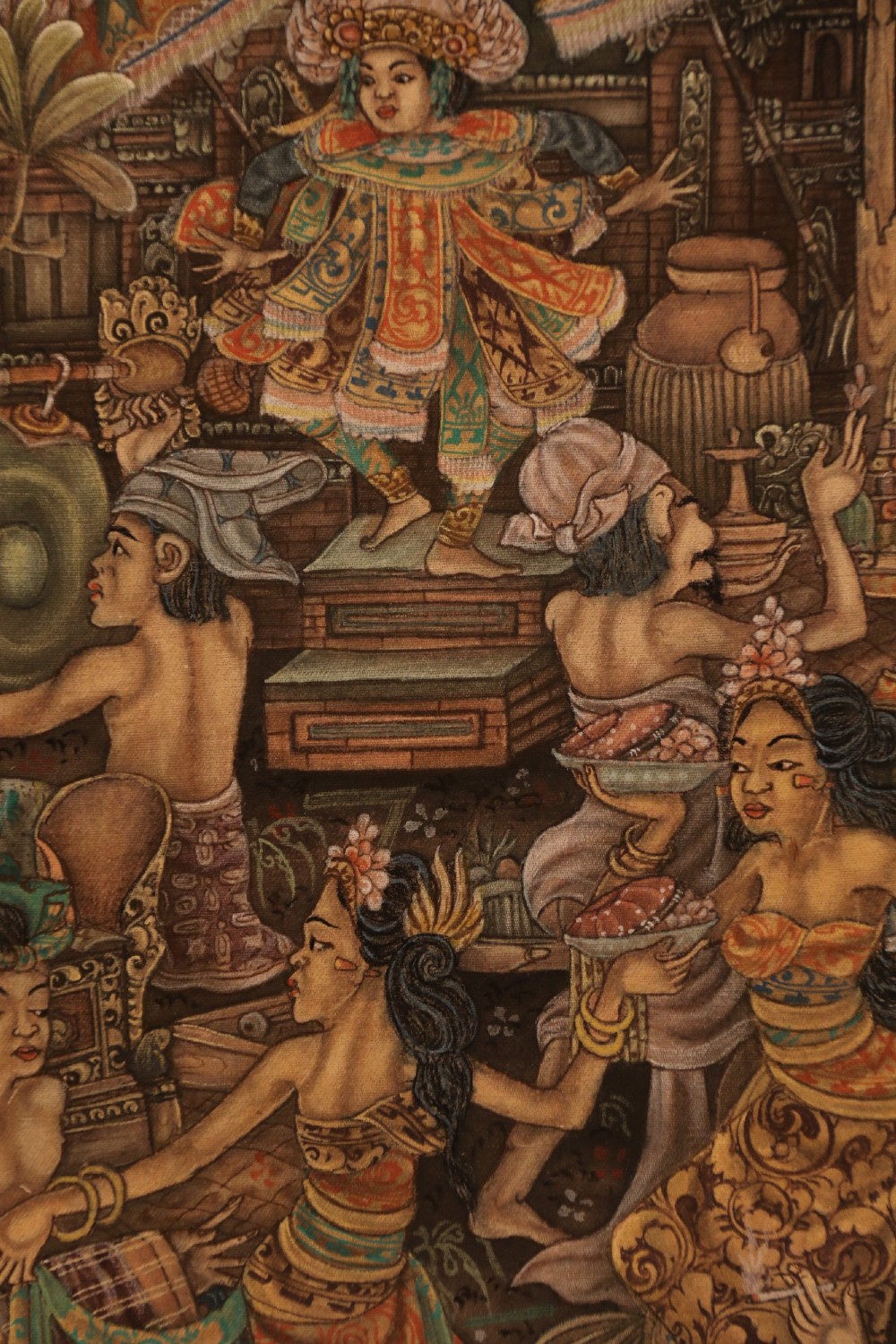 A detailed Balinese painting depicting figures dancing and playing gamelan instruments within a lush landscape, set inside a carved dark wood frame. View XII
