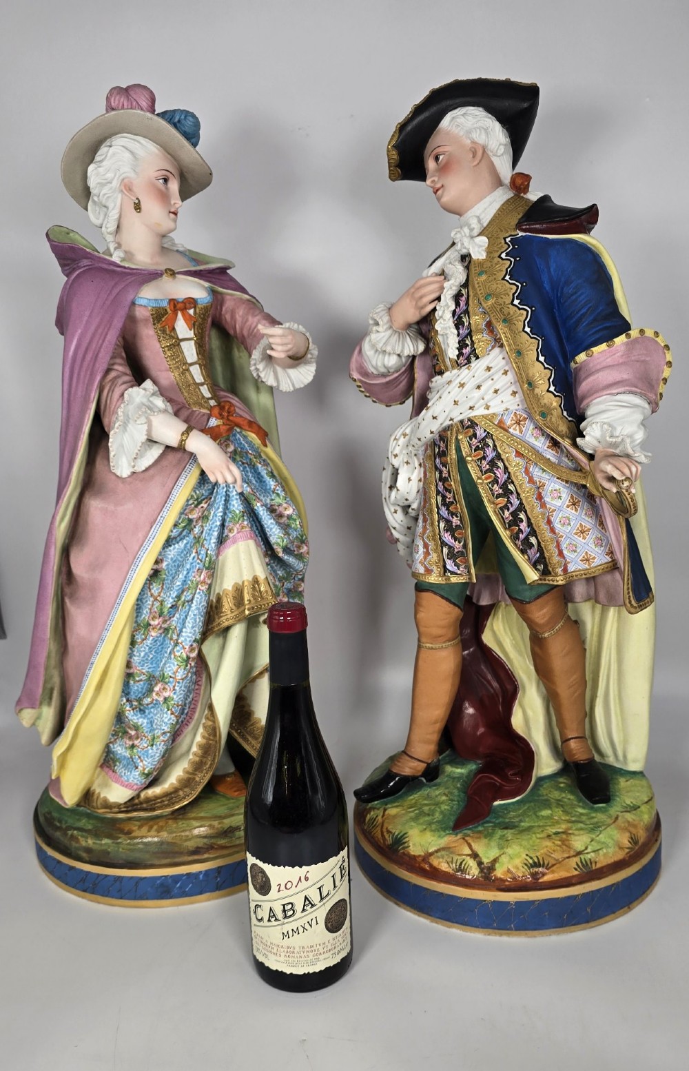 A pair of large hand-painted French porcelain statuettes depicting an 18th-century man and woman in elaborate court attire standing on decorated round pedestals.
