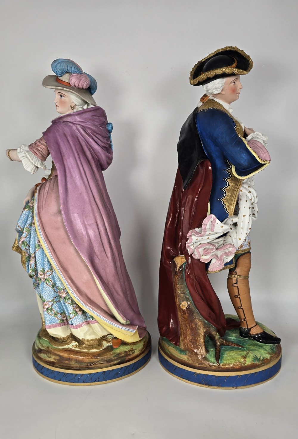 A pair of large hand-painted French porcelain statuettes depicting an 18th-century man and woman in elaborate court attire standing on decorated round pedestals. View II
