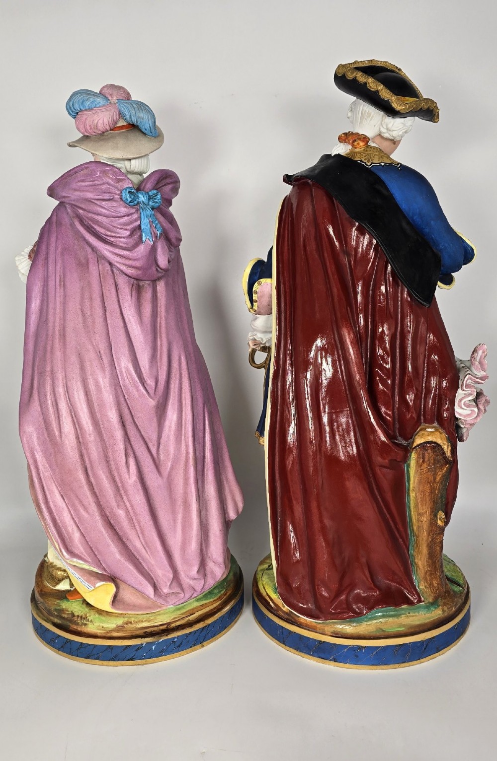 A pair of large hand-painted French porcelain statuettes depicting an 18th-century man and woman in elaborate court attire standing on decorated round pedestals. View III