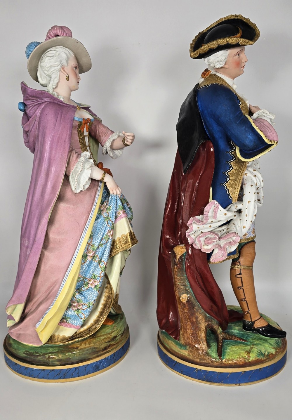 A pair of large hand-painted French porcelain statuettes depicting an 18th-century man and woman in elaborate court attire standing on decorated round pedestals. View IV