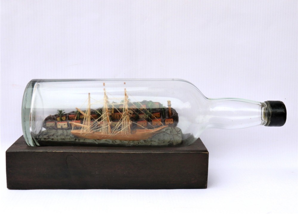 An antique ship in a bottle diorama featuring a detailed wooden sailing vessel and coastal background set on a dark rectangular wooden plinth.