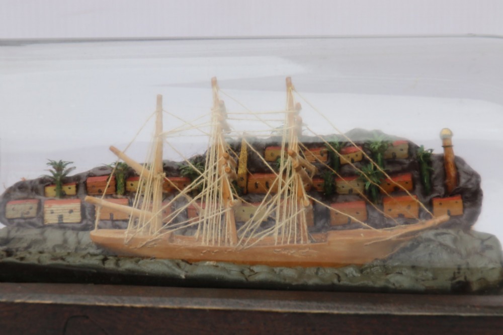 An antique ship in a bottle diorama featuring a detailed wooden sailing vessel and coastal background set on a dark rectangular wooden plinth. View II
