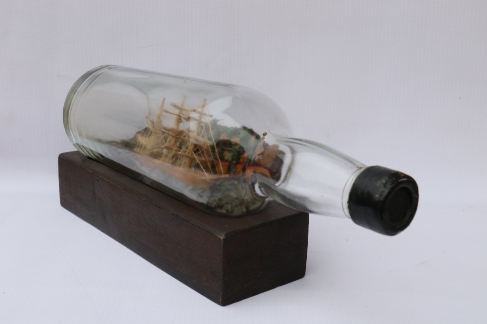 An antique ship in a bottle diorama featuring a detailed wooden sailing vessel and coastal background set on a dark rectangular wooden plinth. View III