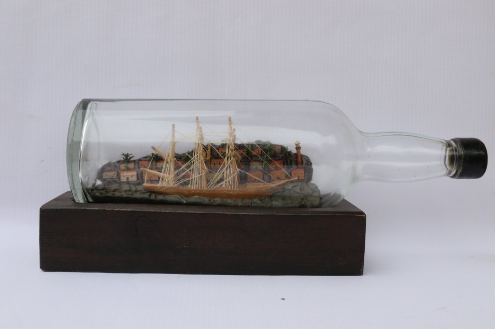 An antique ship in a bottle diorama featuring a detailed wooden sailing vessel and coastal background set on a dark rectangular wooden plinth. View IV