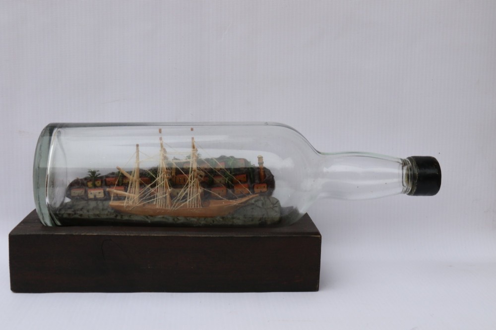 An antique ship in a bottle diorama featuring a detailed wooden sailing vessel and coastal background set on a dark rectangular wooden plinth. View V