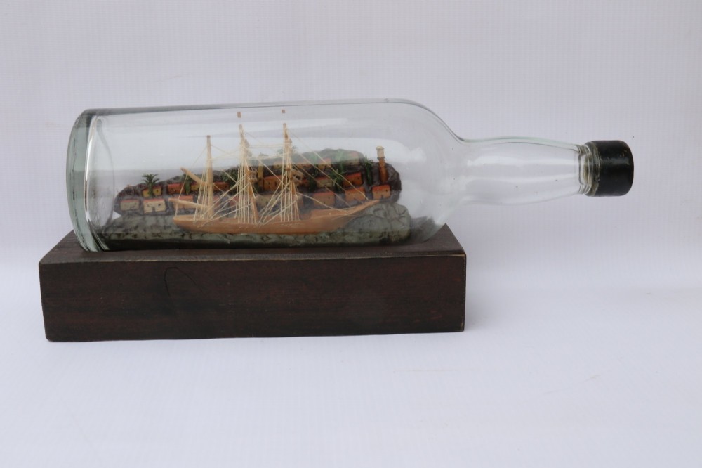 An antique ship in a bottle diorama featuring a detailed wooden sailing vessel and coastal background set on a dark rectangular wooden plinth. View VI