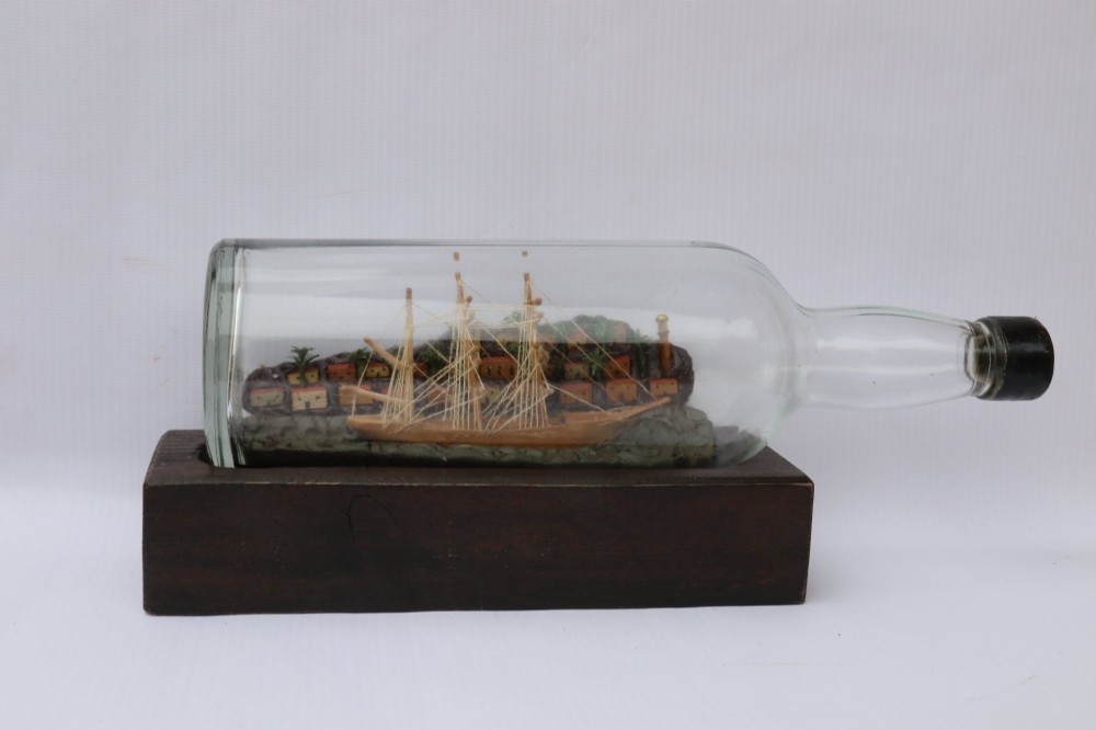 An antique ship in a bottle diorama featuring a detailed wooden sailing vessel and coastal background set on a dark rectangular wooden plinth. View VII