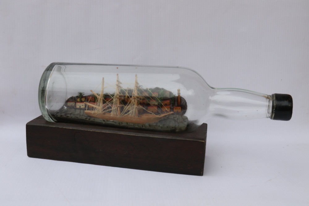 An antique ship in a bottle diorama featuring a detailed wooden sailing vessel and coastal background set on a dark rectangular wooden plinth. View VIII