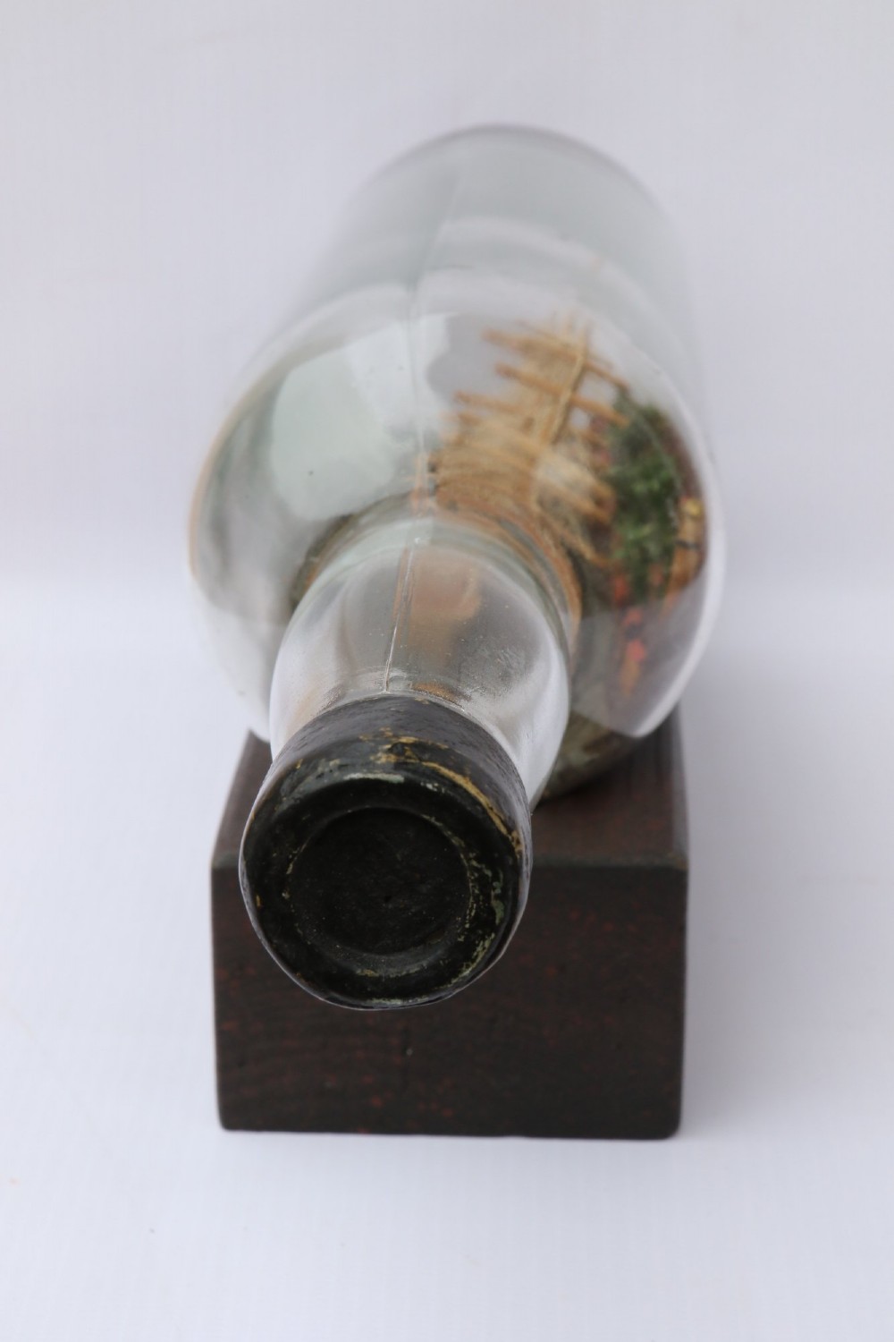 An antique ship in a bottle diorama featuring a detailed wooden sailing vessel and coastal background set on a dark rectangular wooden plinth. View IX