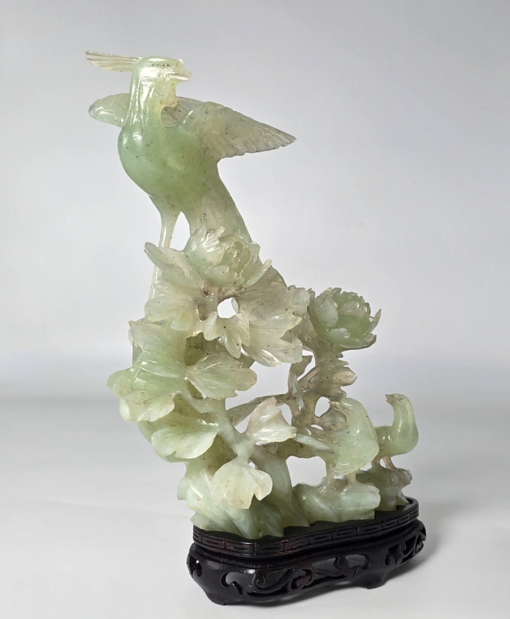 A pale green carved stone sculpture depicting two birds among floral blooms set on a dark carved wood base.