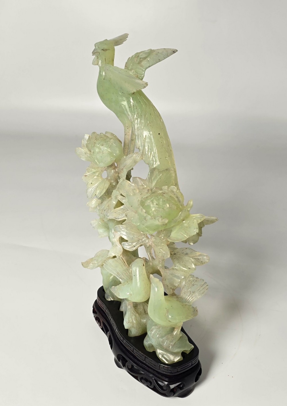 A pale green carved stone sculpture depicting two birds among floral blooms set on a dark carved wood base. View II