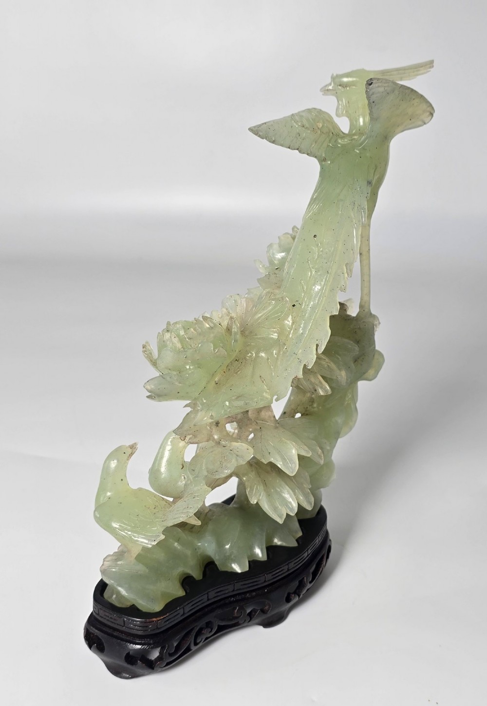 A pale green carved stone sculpture depicting two birds among floral blooms set on a dark carved wood base. View III