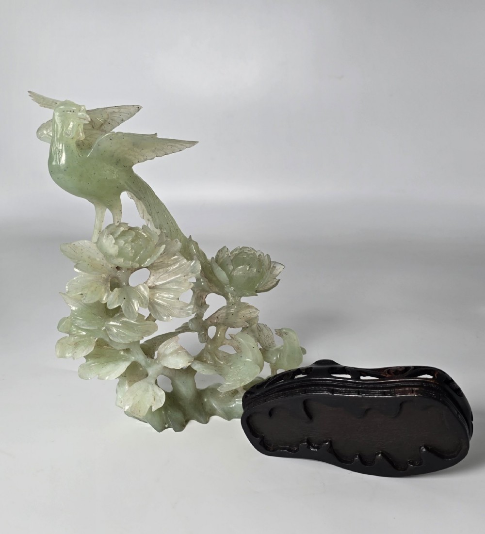 A pale green carved stone sculpture depicting two birds among floral blooms set on a dark carved wood base. View X