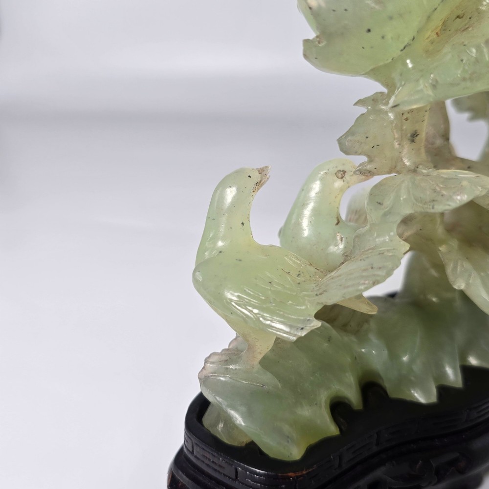A pale green carved stone sculpture depicting two birds among floral blooms set on a dark carved wood base. View XV