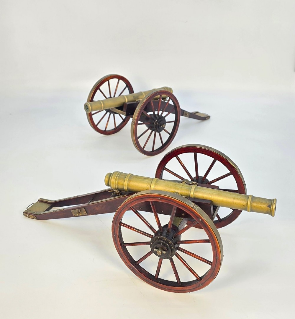 A pair of 1920s model field cannons featuring cast brass barrels mounted on dark wooden spoked-wheel carriages.