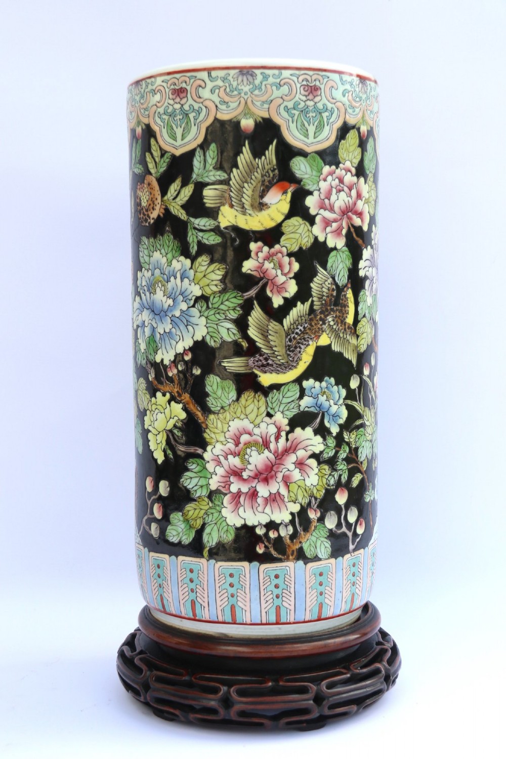 A cylindrical Chinese famille noire porcelain vase decorated with colourful birds and peony flowers set against a black ground, resting on a carved dark hardwood stand.