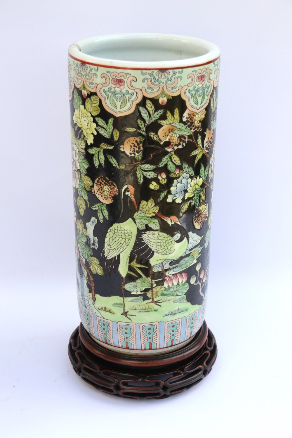 A cylindrical Chinese famille noire porcelain vase decorated with colourful birds and peony flowers set against a black ground, resting on a carved dark hardwood stand. View II