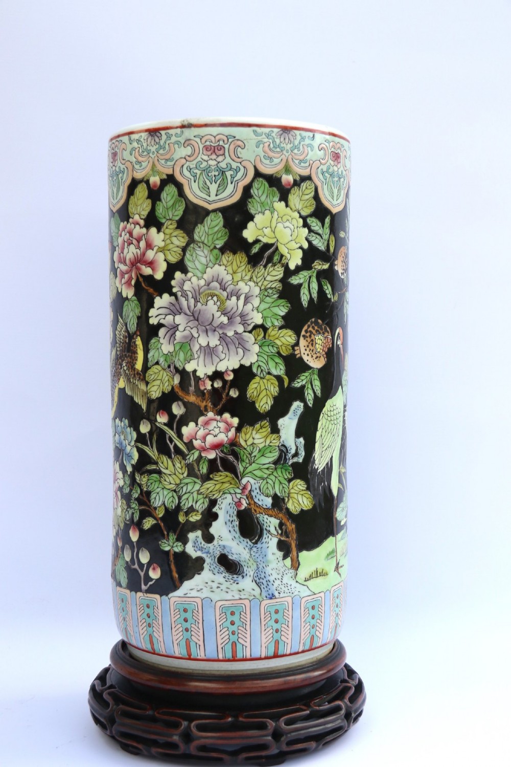 A cylindrical Chinese famille noire porcelain vase decorated with colourful birds and peony flowers set against a black ground, resting on a carved dark hardwood stand. View IV