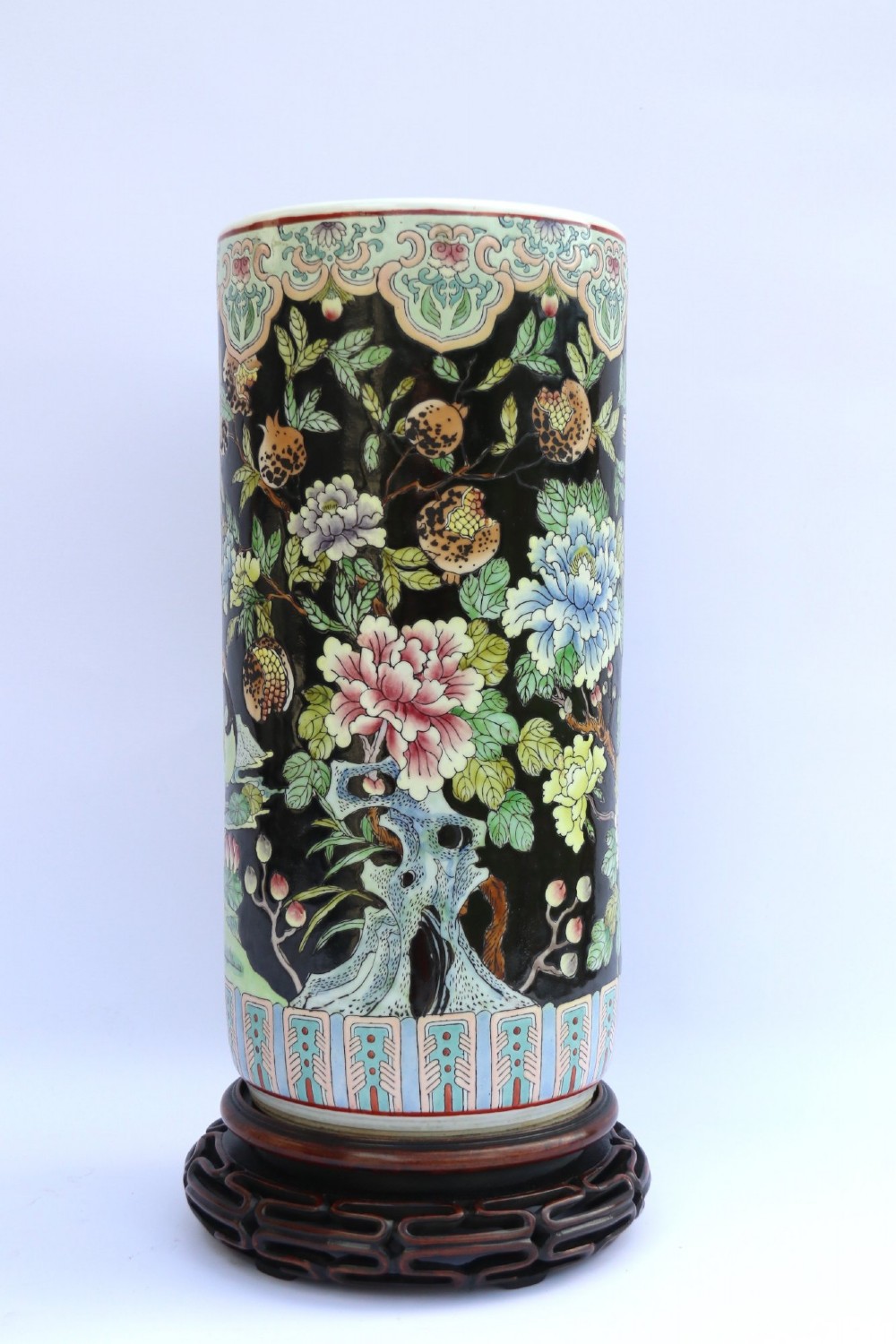 A cylindrical Chinese famille noire porcelain vase decorated with colourful birds and peony flowers set against a black ground, resting on a carved dark hardwood stand. View V