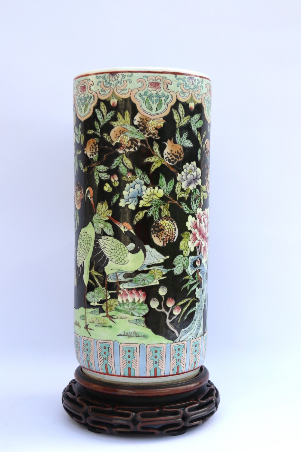 A cylindrical Chinese famille noire porcelain vase decorated with colourful birds and peony flowers set against a black ground, resting on a carved dark hardwood stand. View VI