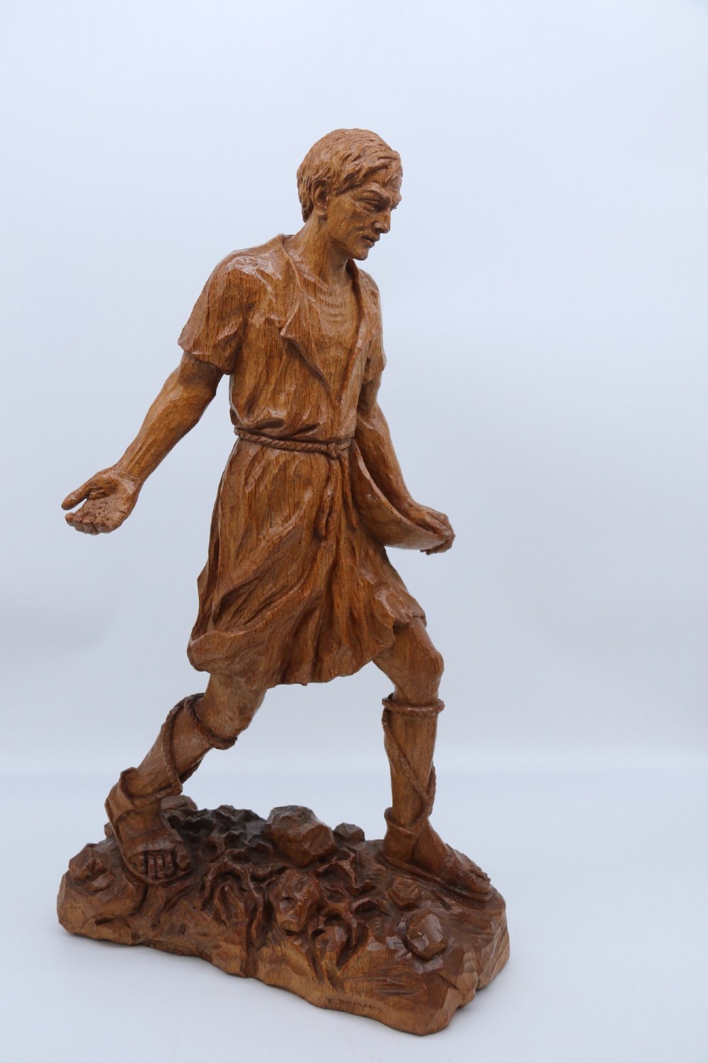 A hand-carved golden oak wooden sculpture of a male farm worker standing on a carved naturalistic base while holding a shallow tray. View II