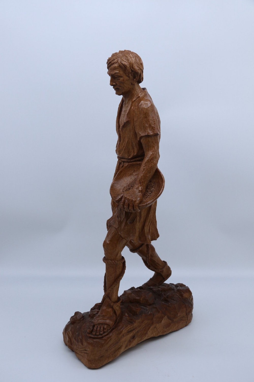 A hand-carved golden oak wooden sculpture of a male farm worker standing on a carved naturalistic base while holding a shallow tray. View VIII