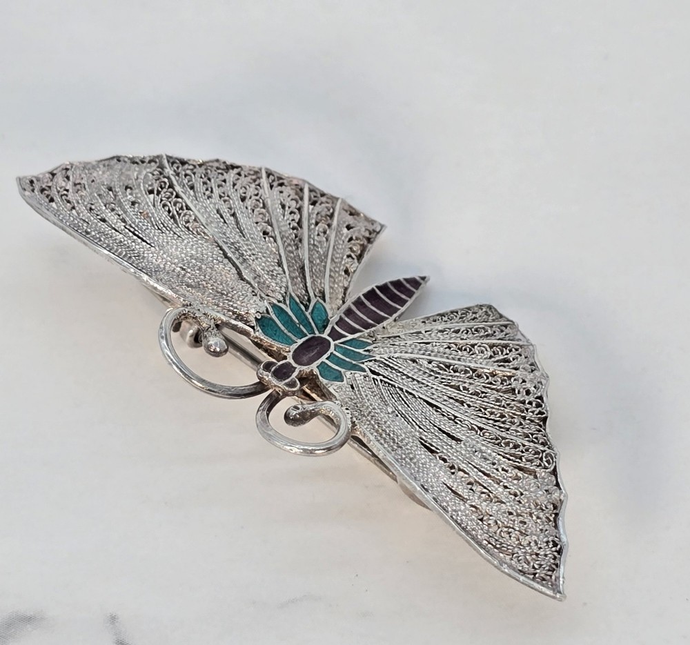 A silver filigree butterfly brooch featuring teal and purple enamelled body details. View VI