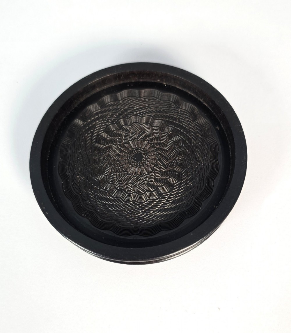 A circular black ebony snuff box with intricate engine-turned geometric patterns and raised beaded details on the lid. View X