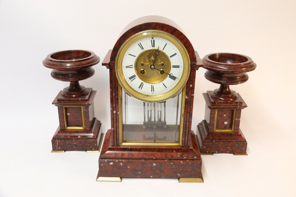 A late nineteenth-century French clock garniture in mottled red marble and gilt brass, featuring a central arch-topped mantel clock with a visible pendulum and two flanking urn cassolettes.