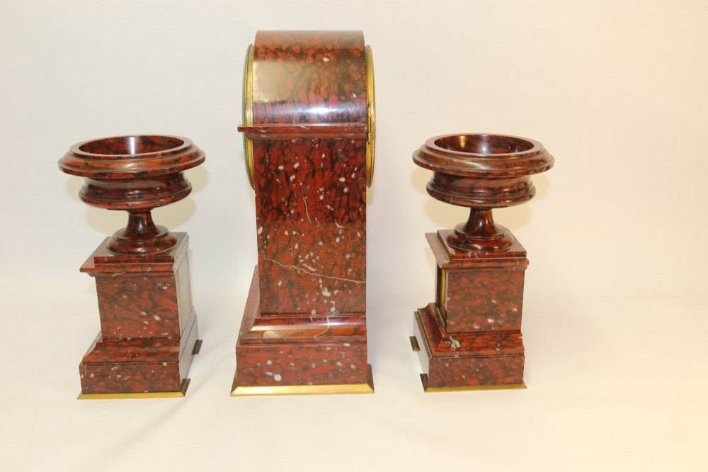 A late nineteenth-century French clock garniture in mottled red marble and gilt brass, featuring a central arch-topped mantel clock with a visible pendulum and two flanking urn cassolettes. View VIII
