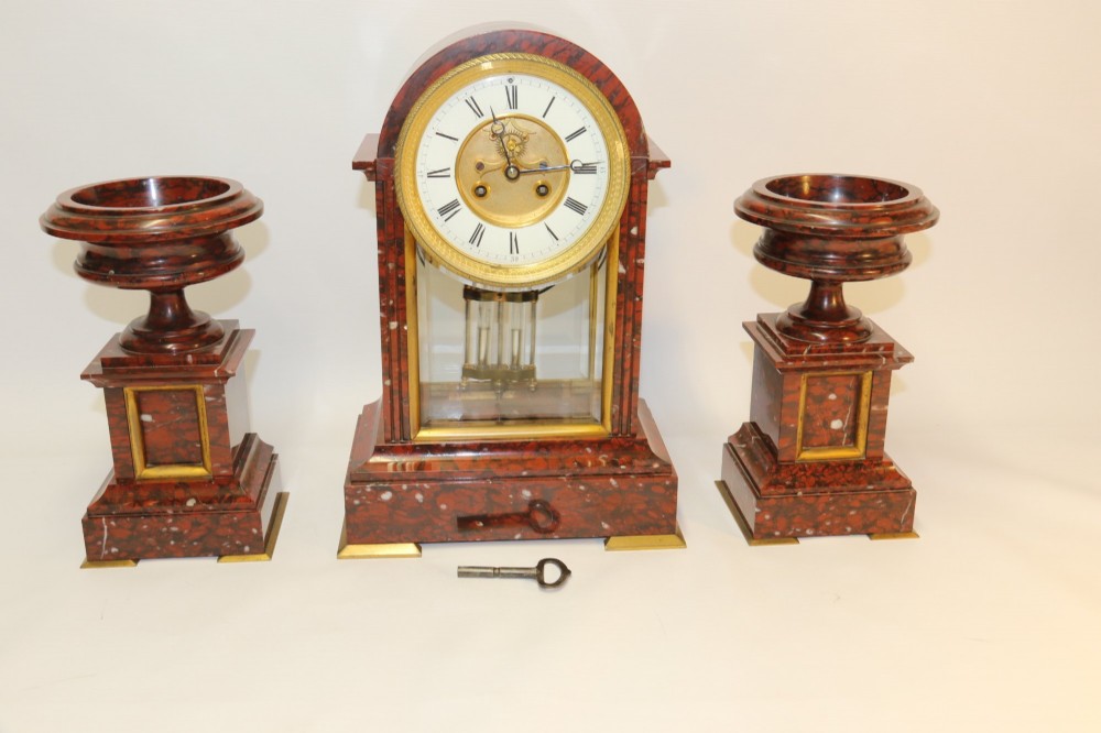 A late nineteenth-century French clock garniture in mottled red marble and gilt brass, featuring a central arch-topped mantel clock with a visible pendulum and two flanking urn cassolettes. View IX