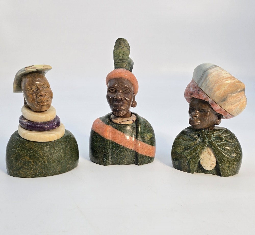 A trio of hand-carved African portrait busts crafted from multi-coloured polished stone depicting figures with distinctive headwear and detailed neck rings.