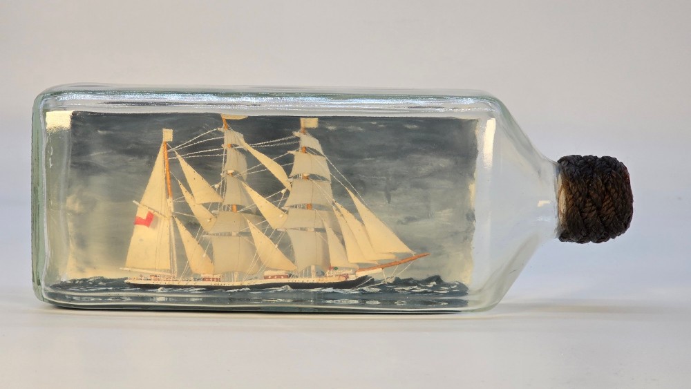 A horizontal clear glass bottle containing a detailed miniature three-masted sailing ship diorama set upon painted blue ocean waves with a rope-bound neck stopper.