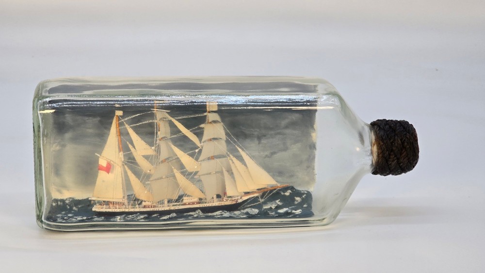 A horizontal clear glass bottle containing a detailed miniature three-masted sailing ship diorama set upon painted blue ocean waves with a rope-bound neck stopper. View II