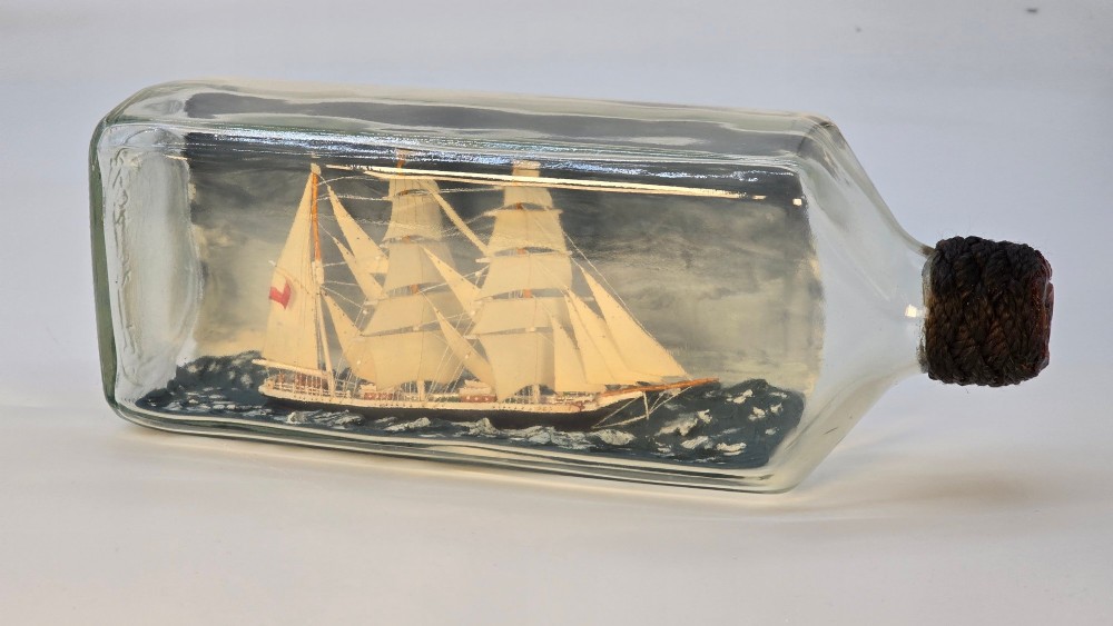 A horizontal clear glass bottle containing a detailed miniature three-masted sailing ship diorama set upon painted blue ocean waves with a rope-bound neck stopper. View III