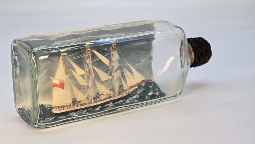 A horizontal clear glass bottle containing a detailed miniature three-masted sailing ship diorama set upon painted blue ocean waves with a rope-bound neck stopper. View IV