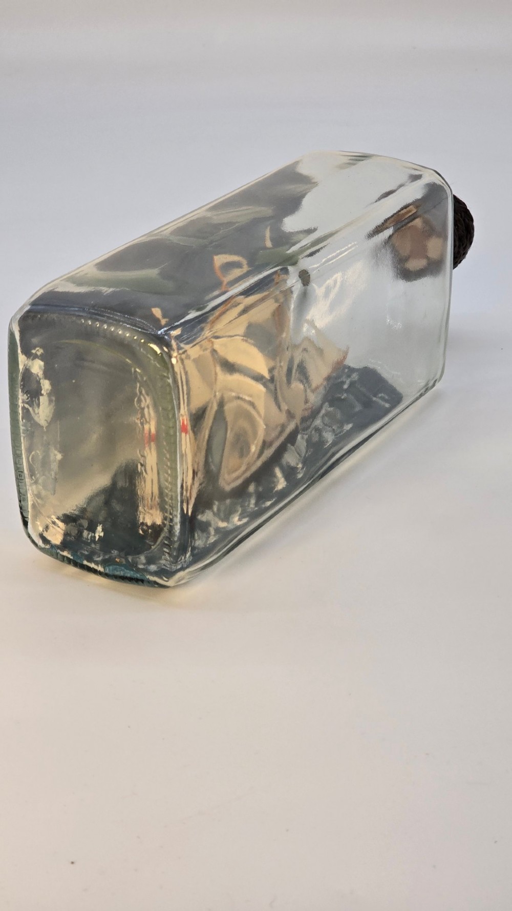 A horizontal clear glass bottle containing a detailed miniature three-masted sailing ship diorama set upon painted blue ocean waves with a rope-bound neck stopper. View V