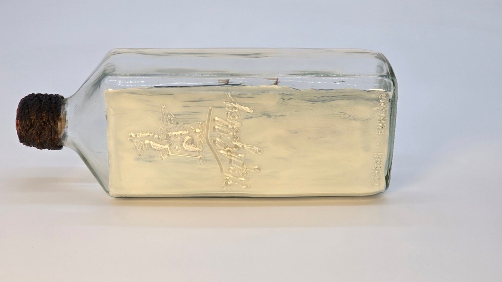 A horizontal clear glass bottle containing a detailed miniature three-masted sailing ship diorama set upon painted blue ocean waves with a rope-bound neck stopper. View VI