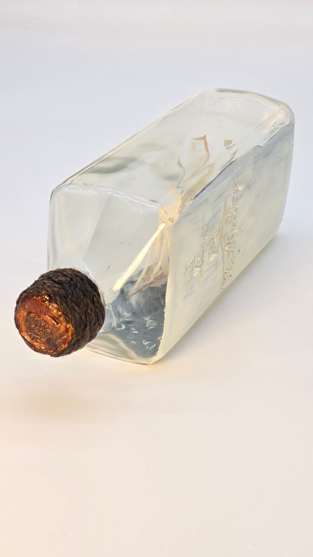 A horizontal clear glass bottle containing a detailed miniature three-masted sailing ship diorama set upon painted blue ocean waves with a rope-bound neck stopper. View VII