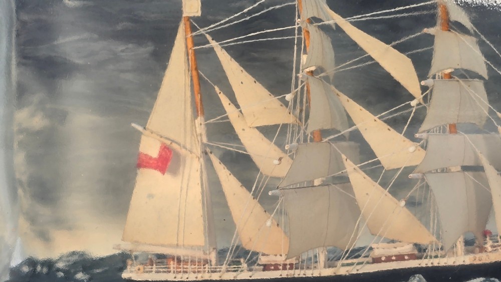 A horizontal clear glass bottle containing a detailed miniature three-masted sailing ship diorama set upon painted blue ocean waves with a rope-bound neck stopper. View X