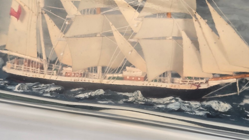 A horizontal clear glass bottle containing a detailed miniature three-masted sailing ship diorama set upon painted blue ocean waves with a rope-bound neck stopper. View XI